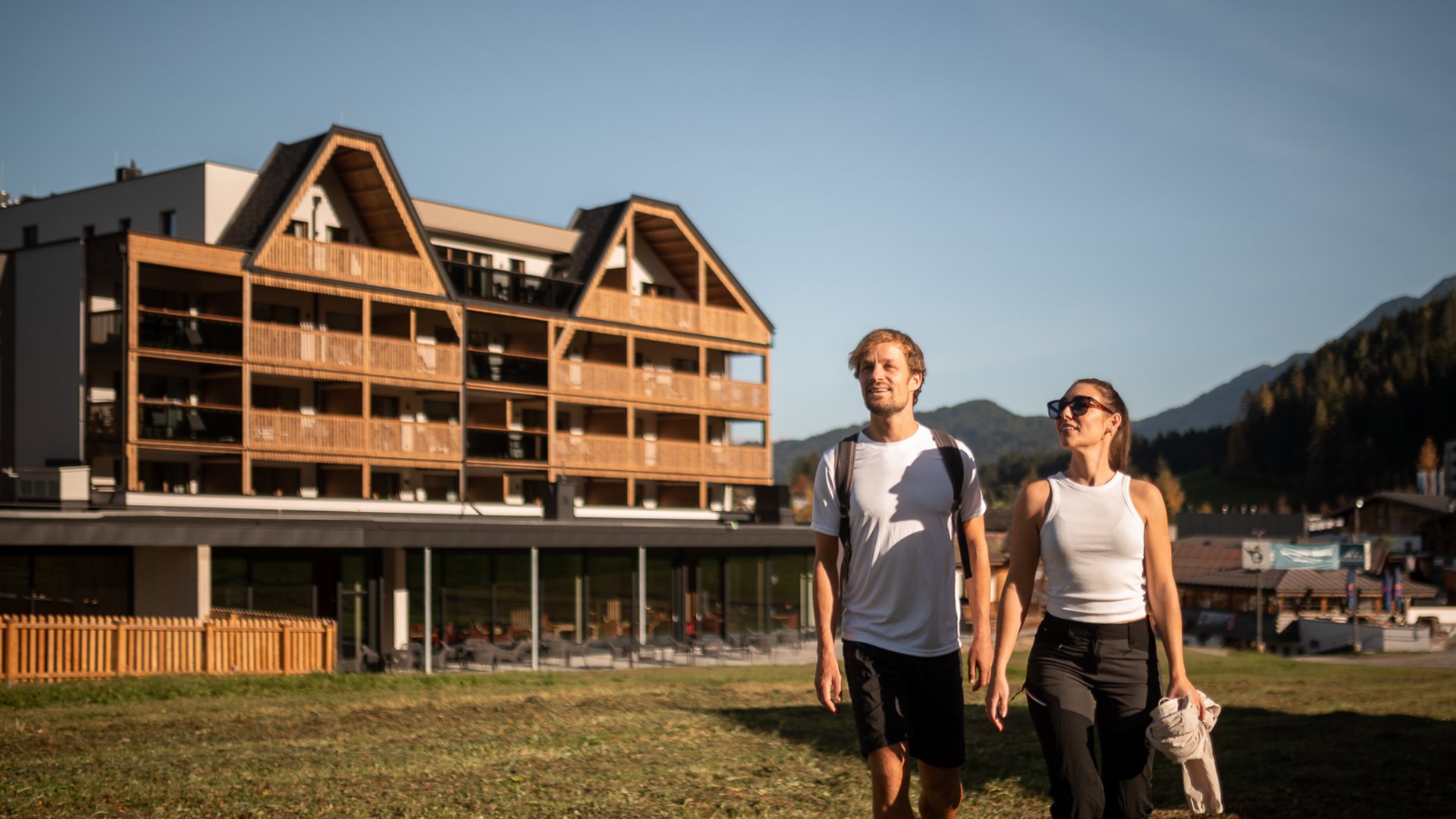 ADEA Lifestyle Suites – romantisches Hotel in Tirol Paar geht im Sonnenschein vor einem Holzhaus in den Bergen spazieren