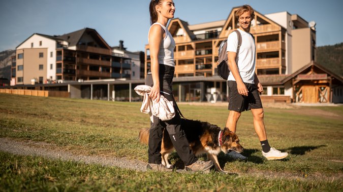ADEA Lifestyle Suites – romantisches Hotel in Tirol Paar geht mit Hund auf Weg vor modernen Chalets spazieren bei Sonnenschein