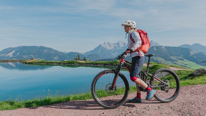 MTB Fieberbrunn – Home of Lässig Frau mit E-Bike auf Bergweg neben Bergsee und Alpenkulisse
