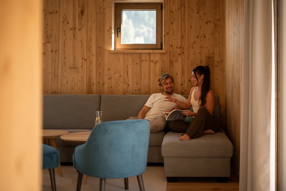 Hochzeitslocation in Tirol: ADEA Lifestyle Suites Paar spricht auf grauem Sofa in einem Raum mit Holzpaneelen