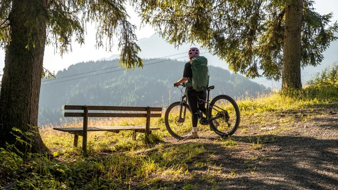 Urlaub für... Fahrradfahrerin mit Helm und Rucksack auf Waldweg neben Bank mit Bergblick