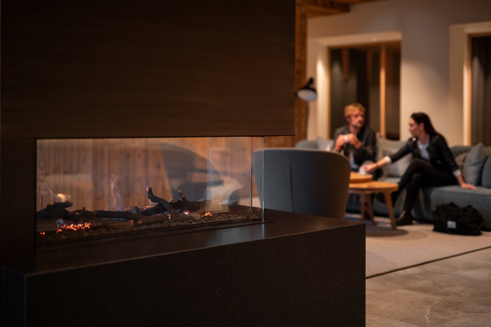 ADEA: dein Hotel mit Fitnessstudio in Österreich Moderner Kamin mit brennenden Holzscheiten und zwei Personen auf dem Sofa im Hintergrund