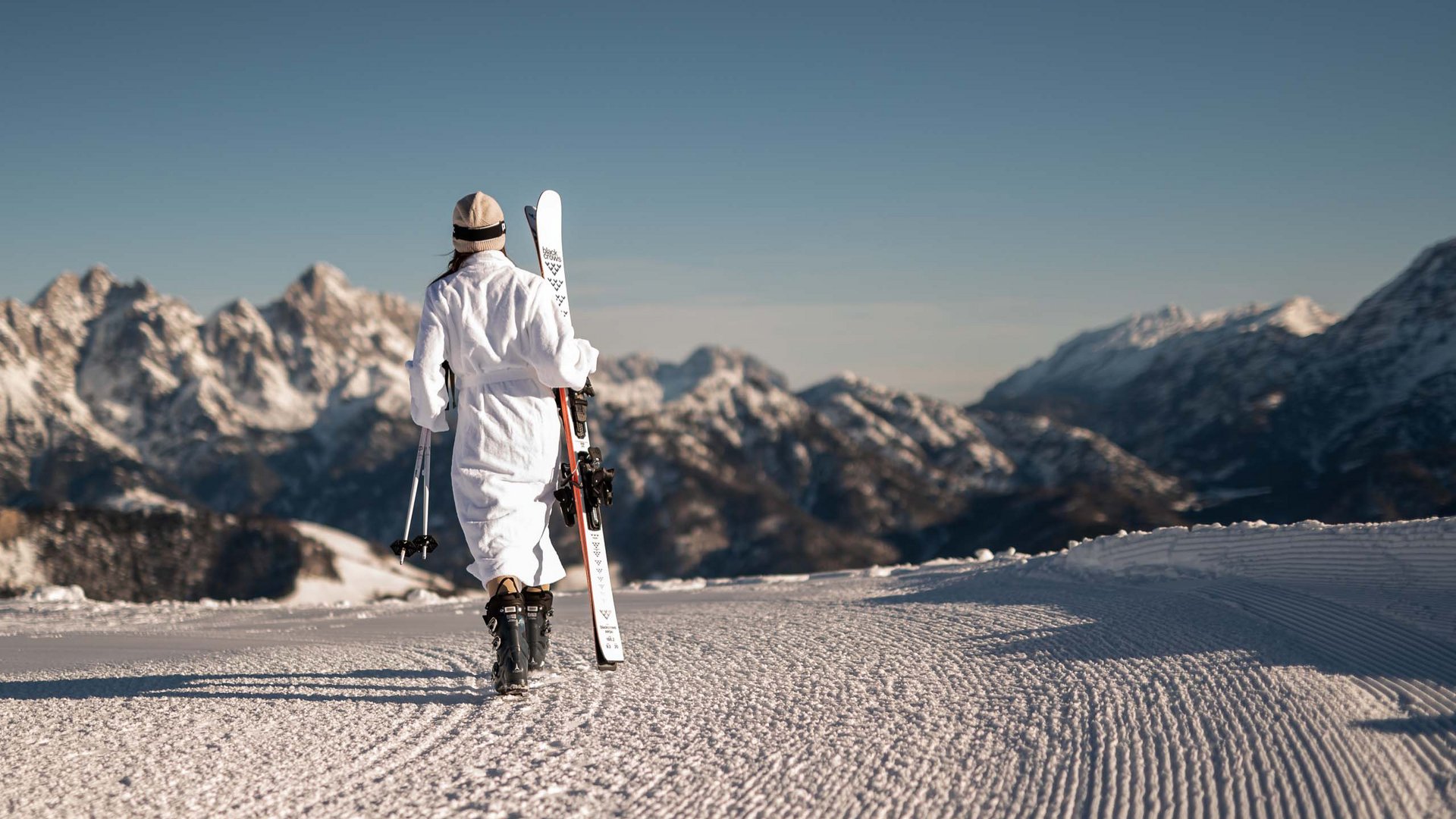 ADEA Lifestyle Suites – romantisches Hotel in Tirol Person im Bademantel trägt Ski und Stöcke auf verschneitem Berg mit Alpen im Hintergrund