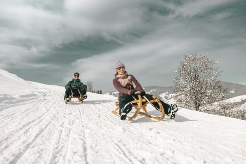 Urlaub für... Zwei Personen rodeln fröhlich auf einem schneebedeckten Hügel im Winter