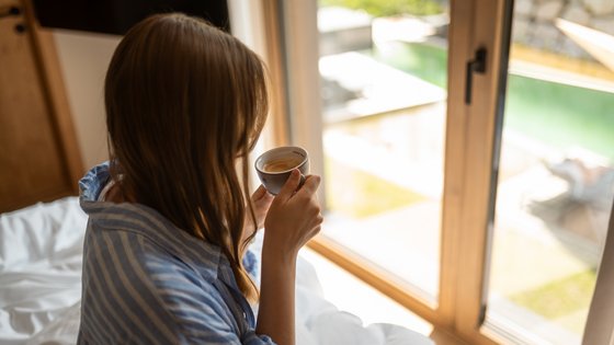 Frau sitzt auf dem Bett mit einer Tasse Kaffee in der Hand und blickt durchs Fenster direkt auf den Schwimmteich Frau sitzt auf dem Bett mit einer Tasse Kaffee in der Hand und blickt durchs Fenster direkt auf den Schwimmteich