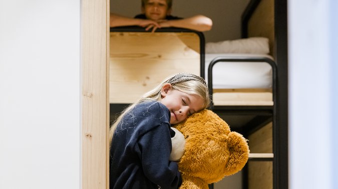 Mädchen hat einen riesigen Teddybär im Arm, im Hintergrund ist ein Stockbett auf dem ein Junge sitzt Mädchen hat einen riesigen Teddybär im Arm, im Hintergrund ist ein Stockbett auf dem ein Junge sitzt