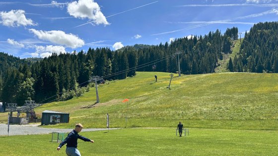 Urlaub für... Mann spielt Fußball auf Wiese vor bewaldetem Hang mit Gondellift