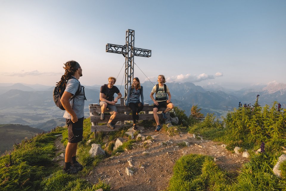 Drinks in Fieberbrunn: Nirgends so gut wie hier! Vier Wanderer auf Berggipfel neben großem Gipfelkreuz bei Sonnenuntergang
