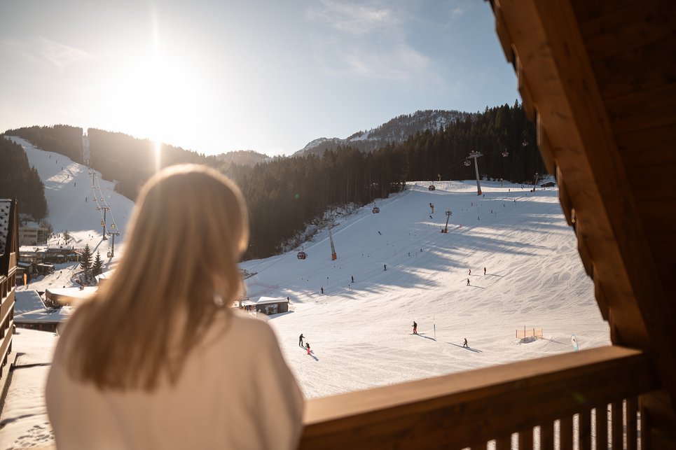Hotel in Fieberbrunn: ADEA Lifestyle Suites Frau blickt von Holzbalkon auf sonnige Skipiste mit Skifahrern und Gondeln