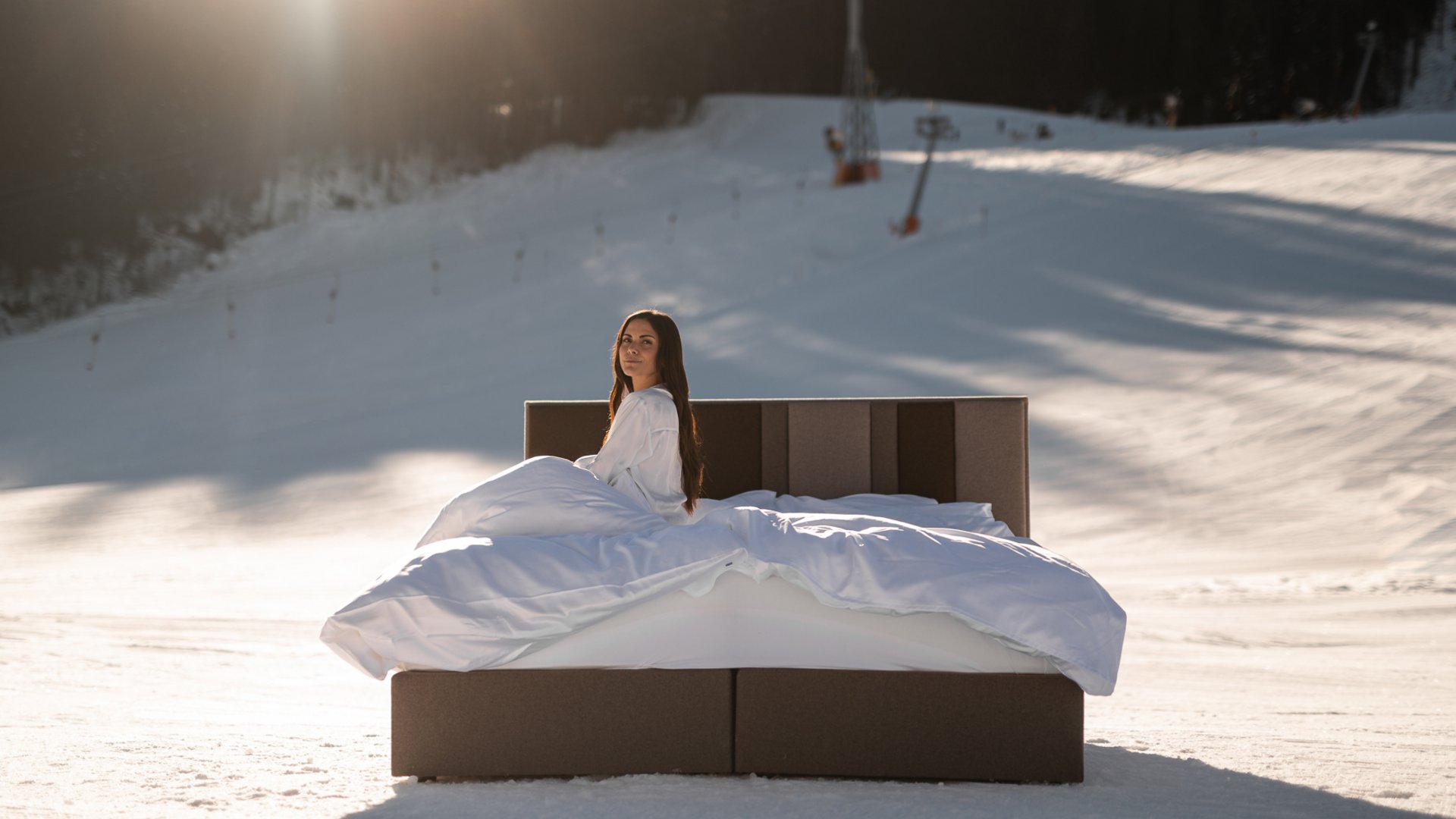 Hotel in Fieberbrunn: ADEA Lifestyle Suites Frau sitzt in Bett mit weißen Decken auf verschneitem Hügel bei Sonnenlicht