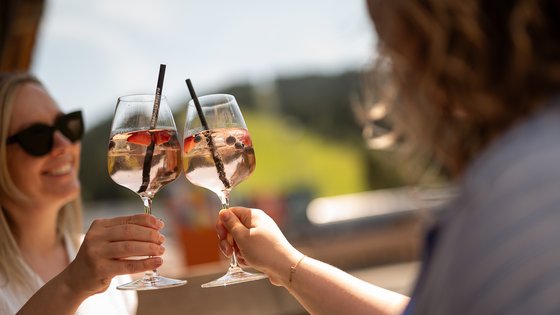 Hotel in Fieberbrunn: ADEA Lifestyle Suites Zwei Frauen stoßen mit Gläsern mit fruchtigem Cocktail an im Sonnenlicht