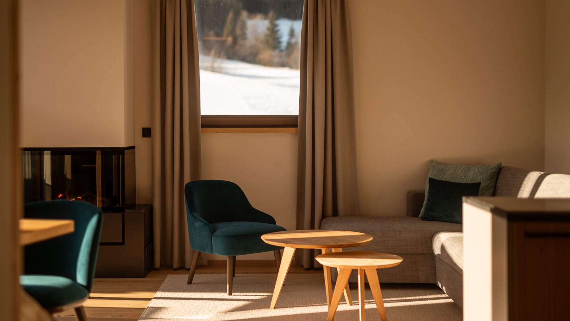 Hotel in Fieberbrunn: ADEA Lifestyle Suites Gemütliches Wohnzimmer mit Kamin, zwei runden Holztischen und Ausblick auf schneebedeckte Landschaft