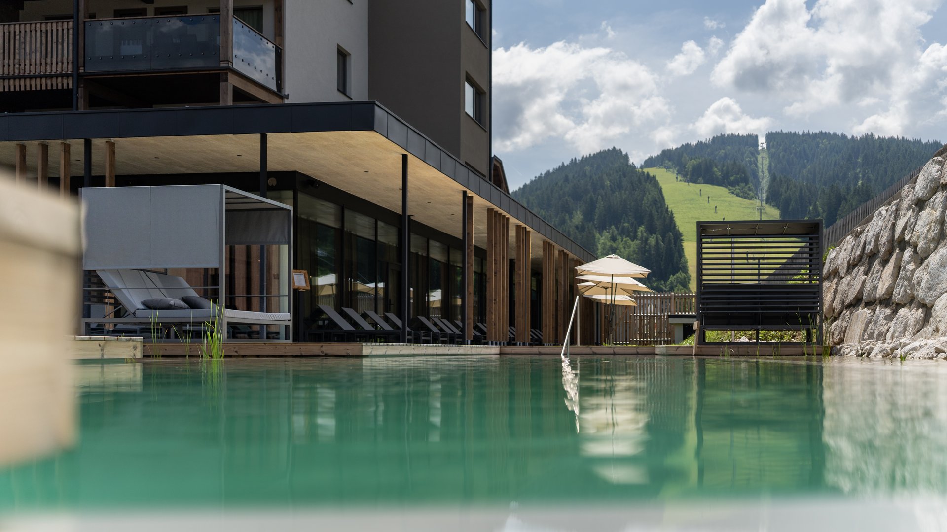Fieberbrunn: Wellnesshotel gesucht? Moderner Hotelpool mit Liegestühlen und Bergblick bei bewölktem Himmel