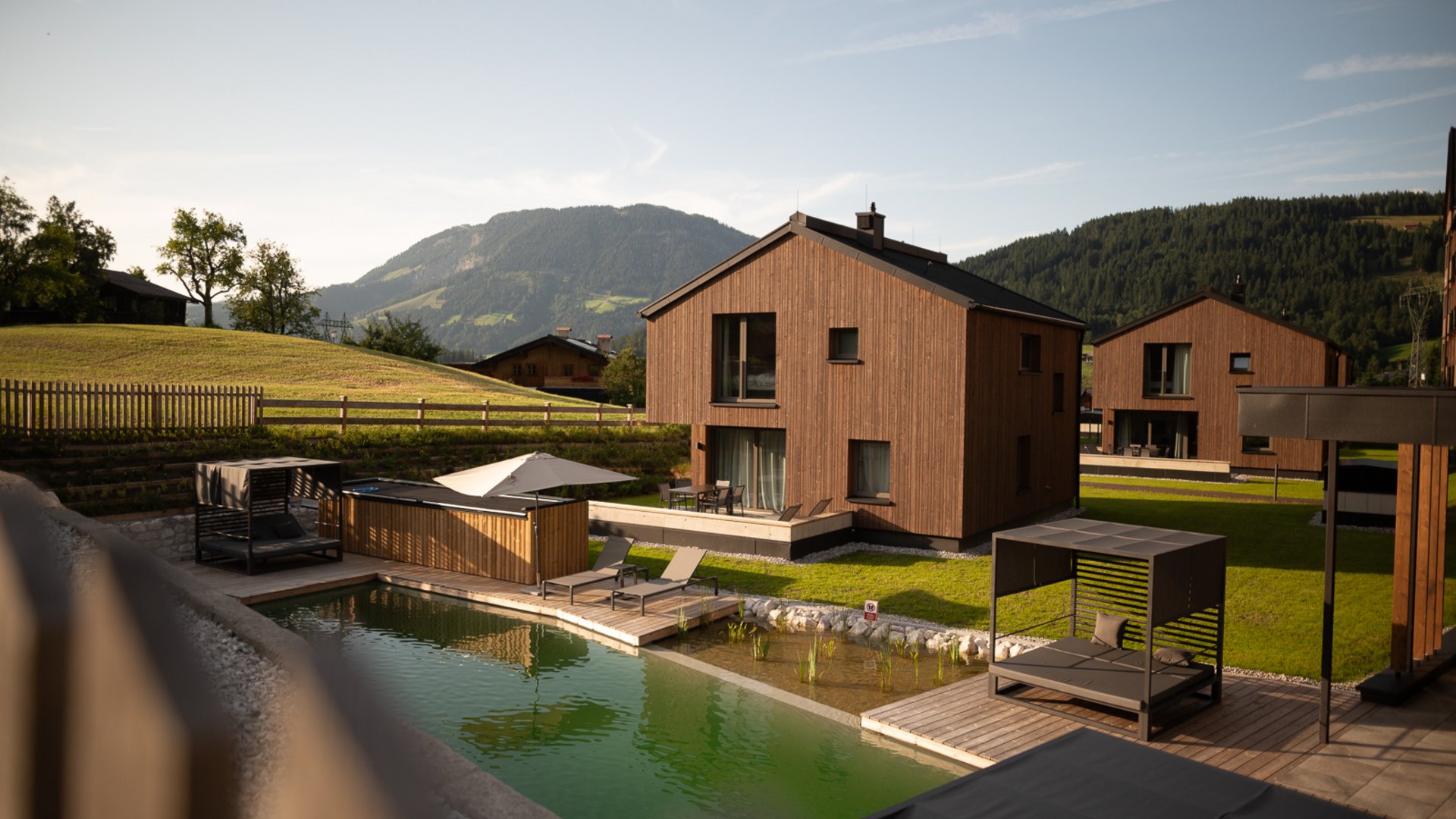 Hotel in Fieberbrunn: ADEA Lifestyle Suites Moderne Holzhäuser mit Liegestühlen und natürlichem Pool in einer Berglandschaft